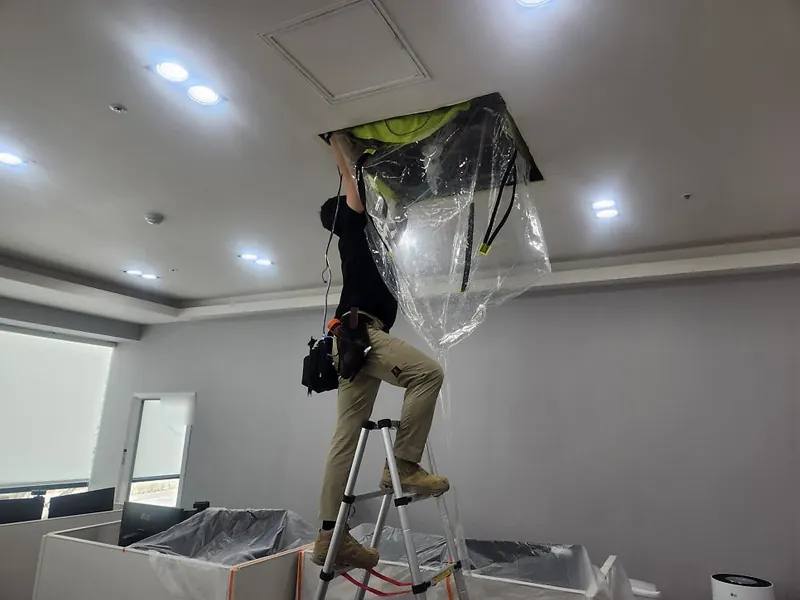 작업자가 사다리 위에서 천장형 에어컨에 방수 가대를 씌우고 아래 집기에 비닐을 덮어 보양하는 모습